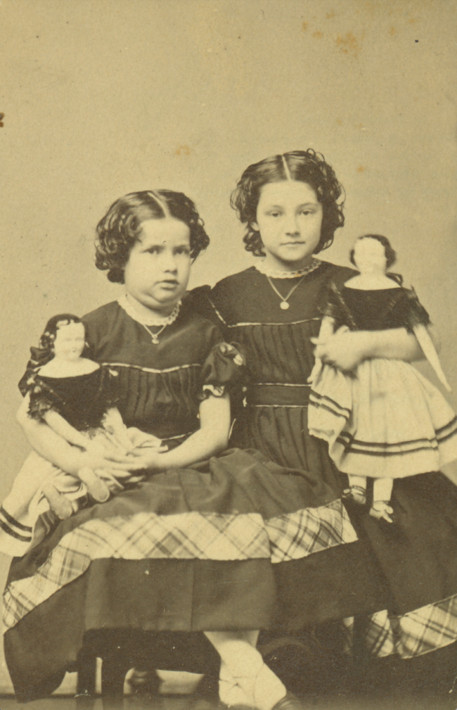 Two young girls in vintage dresses, each holding a doll, with a sepia-tone background.