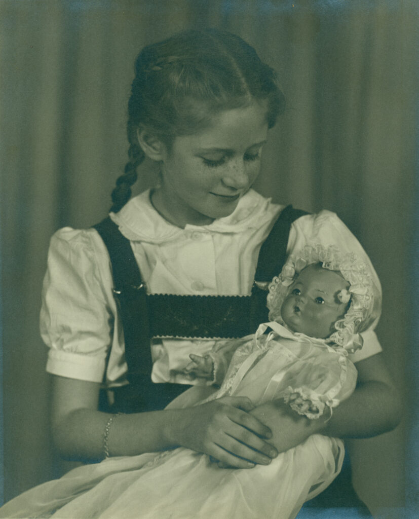 The image depicts a young girl with braided hair, wearing a short-sleeved, white blouse underneath a dark-colored pinafore dress. She is gently cradling a baby doll dressed in a lace-trimmed bonnet and a long, white gown.