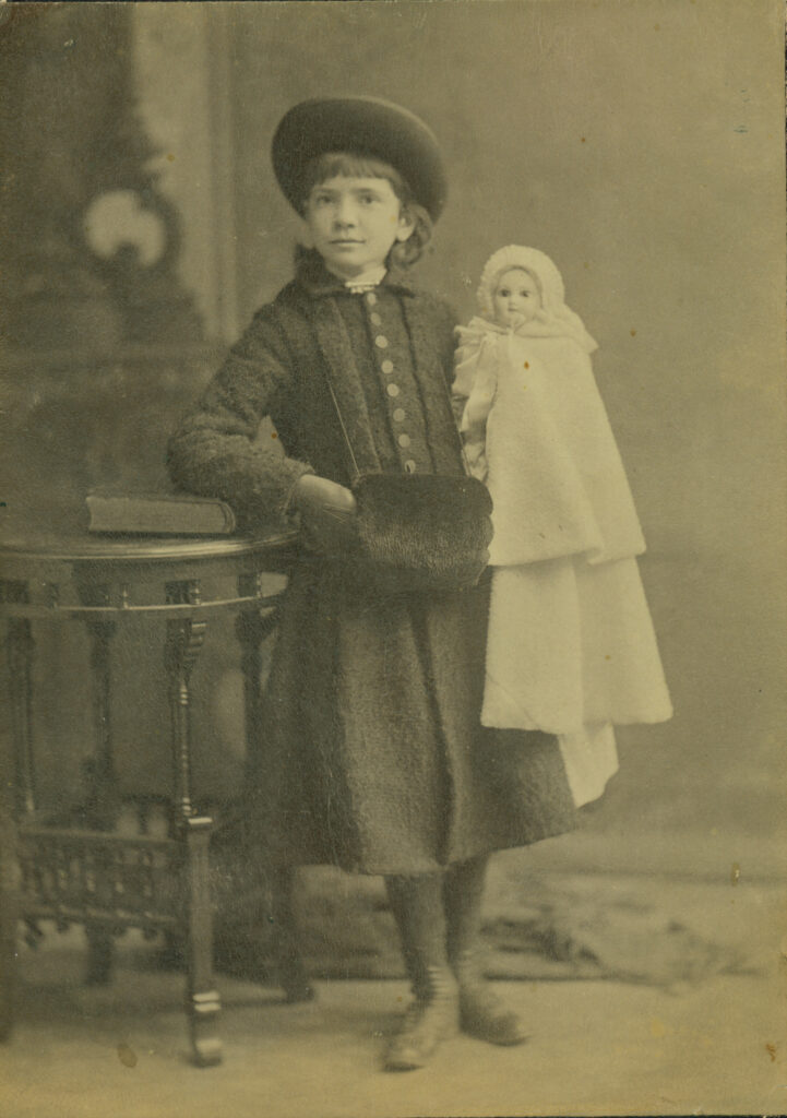 A sepia-toned photo of a child in vintage clothing holding a doll, standing beside an ornate table with a book on it.