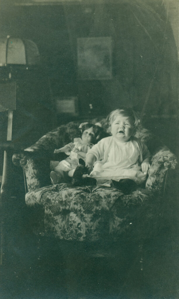 A sepia-toned photograph of a child sitting on a floral-patterned armchair with a doll beside them.