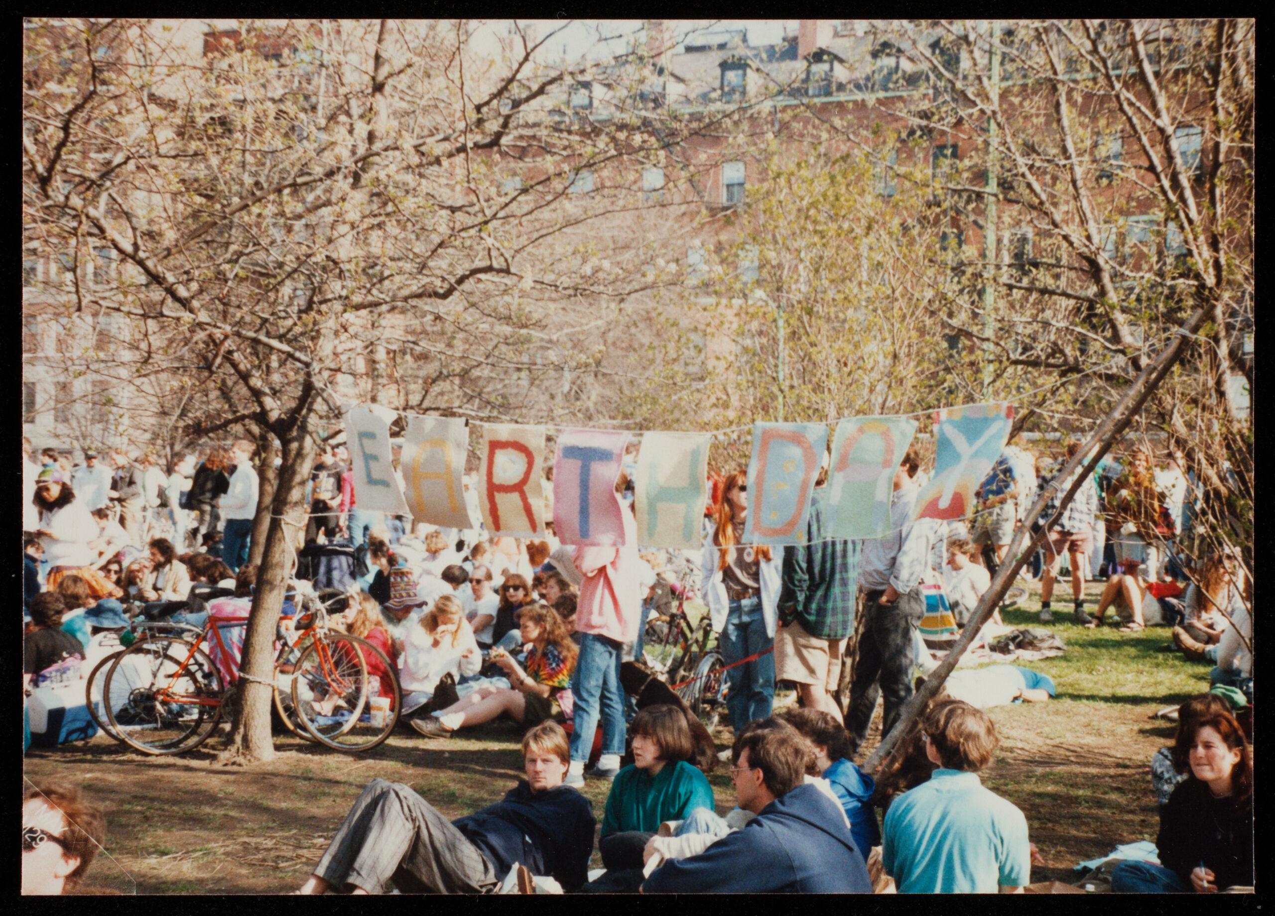 People celebrating Earth Day in a park with a colorful banner and trees in the background. Transcribed Text: EARTH DAY