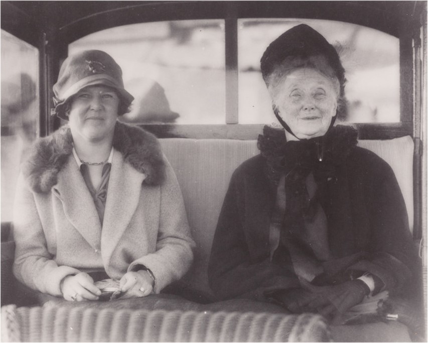 Two women sitting in a carriage, wearing early 20th-century attire, including hats and coats.
