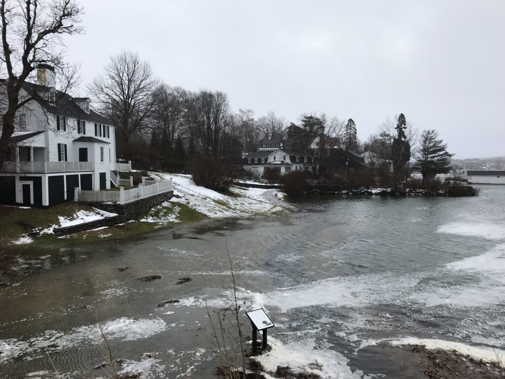 Storm waters surges around white houses on a partially snow-covered embankment.