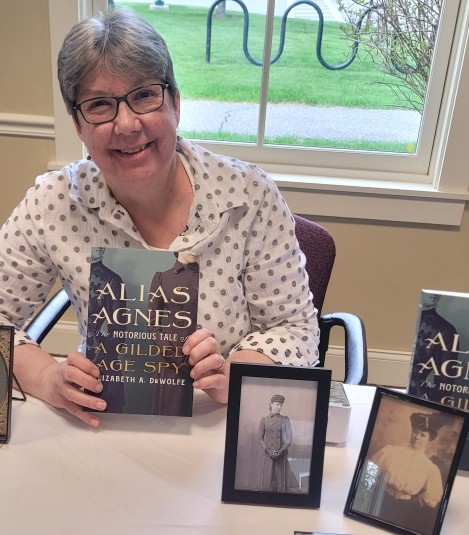 Person holding the book "Alias Agnes" at a table with historical photographs. Transcribed Text: ALIAS AGNES THE NOTORIOUS TALE OF A GILDED AGE SPY ELIZABETH A. DEWOLFE
