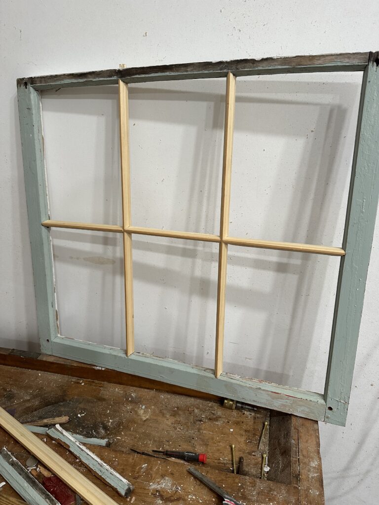 A weathered wooden window frame with six sections, resting on a wooden workbench with scattered tools.