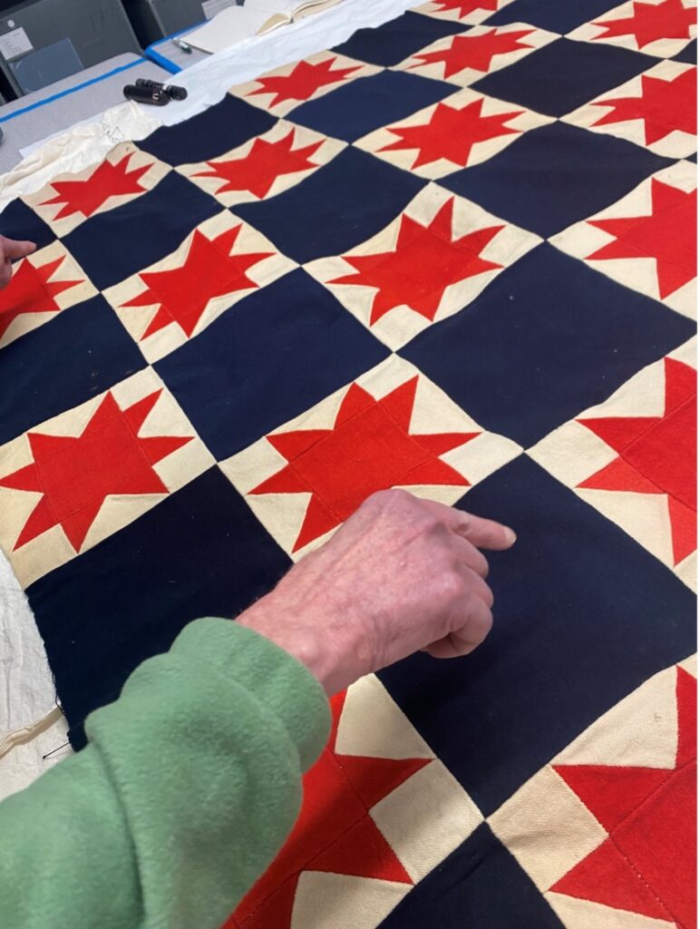 Quilt with red star pattern on blue and cream squares, with a hand pointing at it.