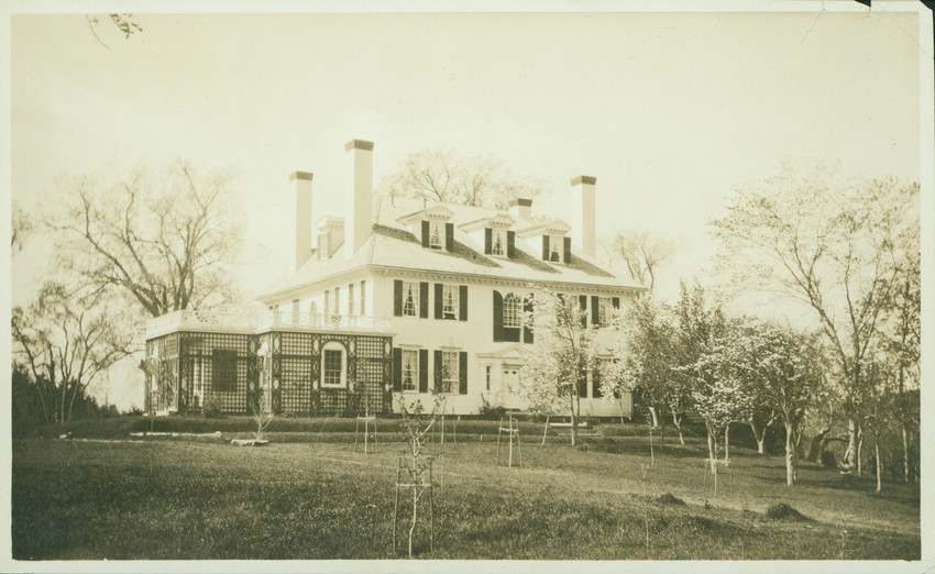 Large colonial-style house with chimneys and a hipped roof surrounded by trees and lawn.