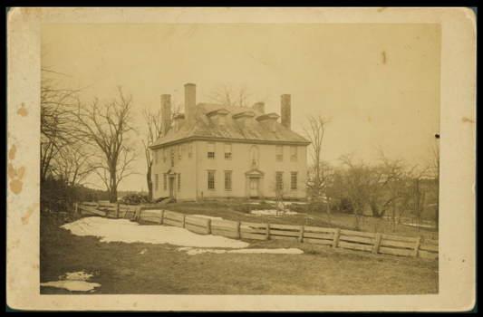 Sepia-toned photograph of a large house with a mansard roof and multiple chimneys, surrounded by barren trees and patches of snow.