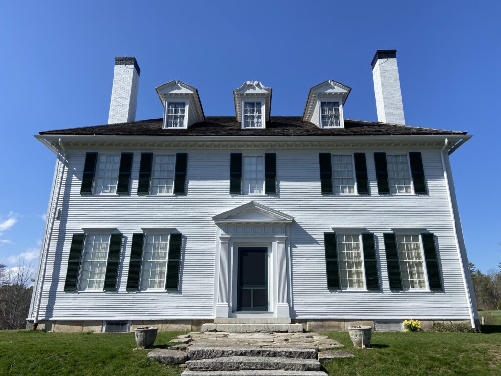 A historic white clapboard house with black shutters and stone steps leading to the entrance, against a clear blue sky.