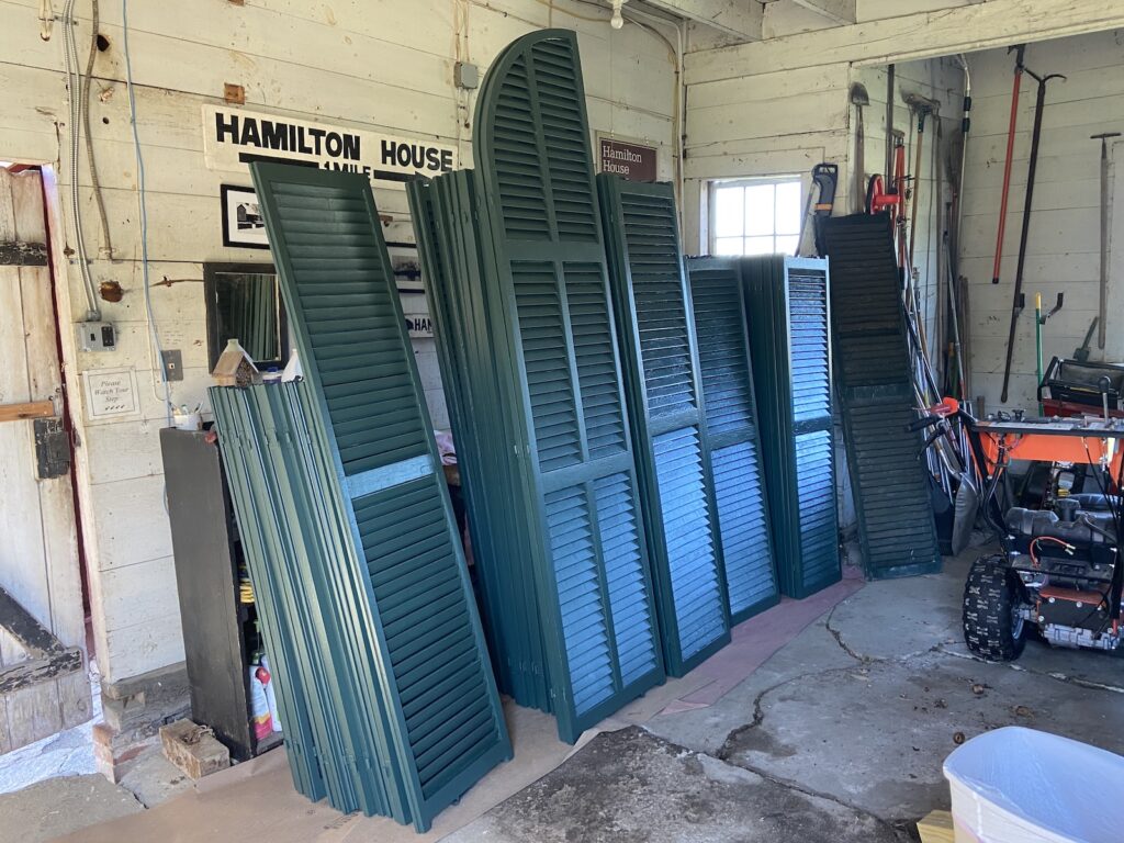 Green wooden shutters stacked in a garage with a sign reading "Hamilton House" on the wall.