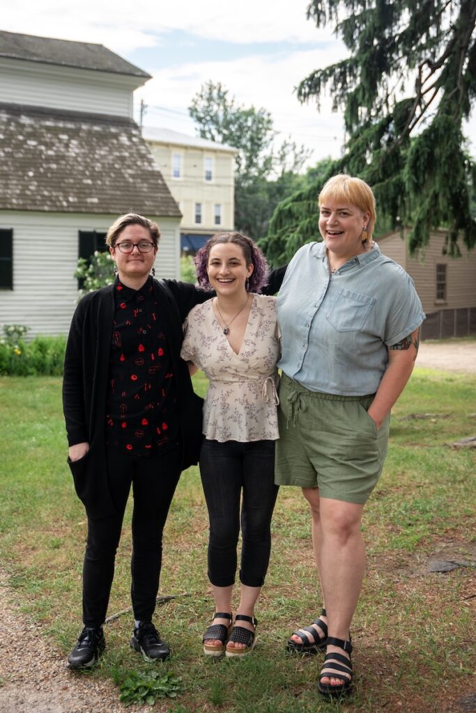 Three people standing outside on grass with a building and trees in the background, smiling with arms around each other.