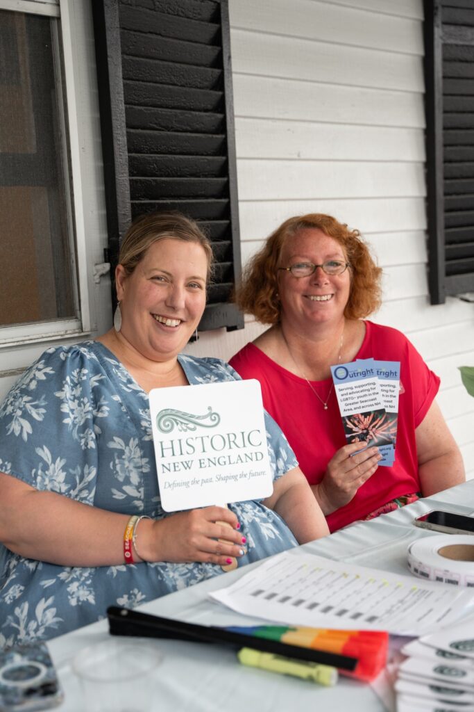 Two women seated at a table holding informational materials, including a fan and a brochure.