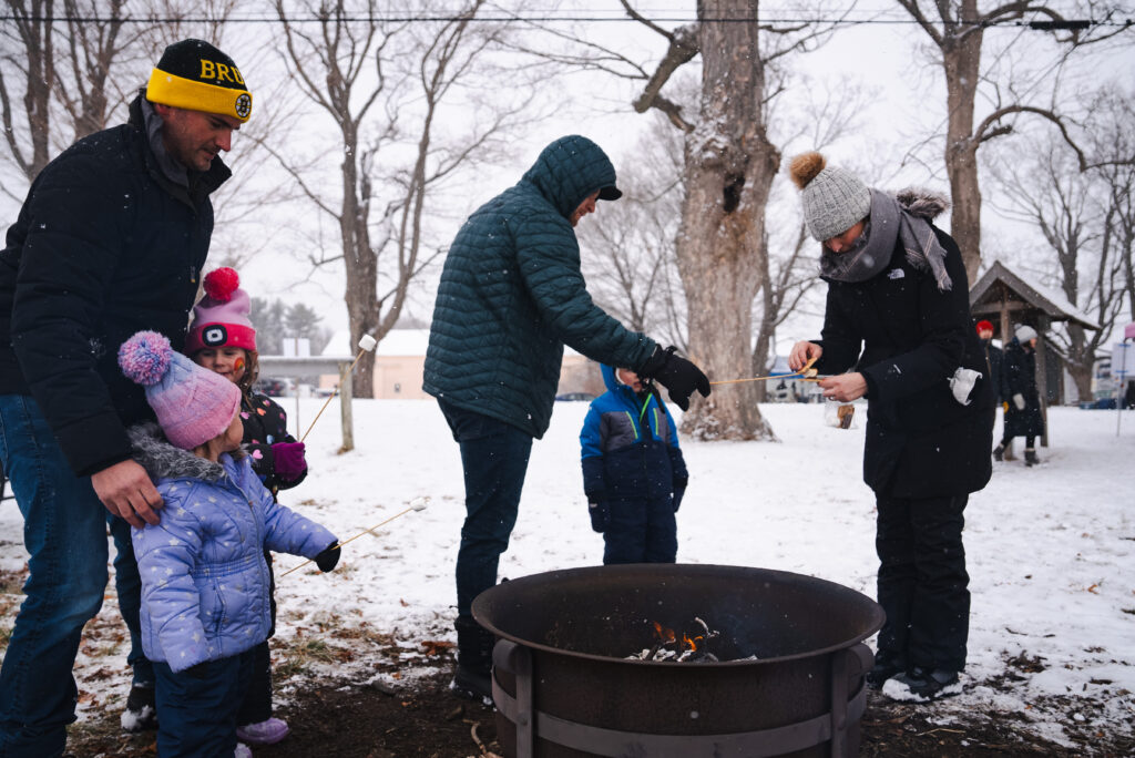 People roasting marshmallows over a fire pit in a snowy open space.