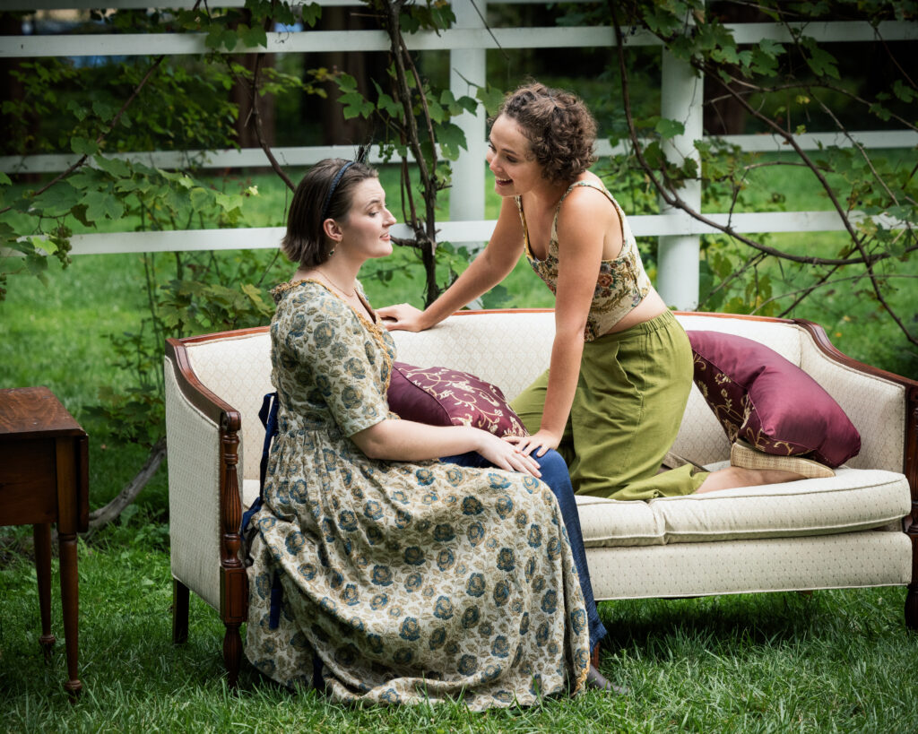 Two women smiling at each other on a vintage sofa with a vine-covered fence in the background.