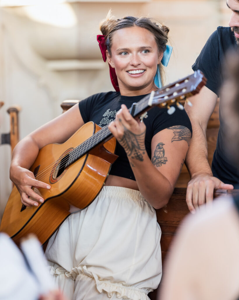 Person playing an acoustic guitar, smiling, with colorful ribbons in their hair.