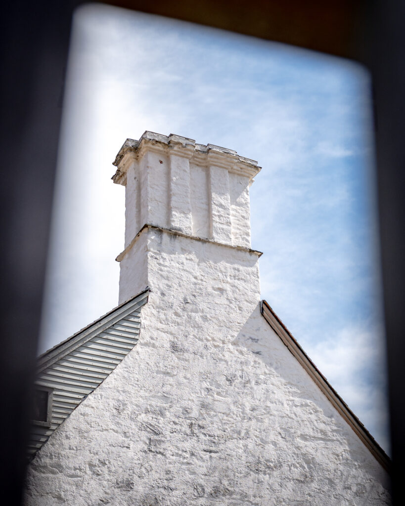 A white brick chimney extends above a historic building facade with gray siding under a blue sky.