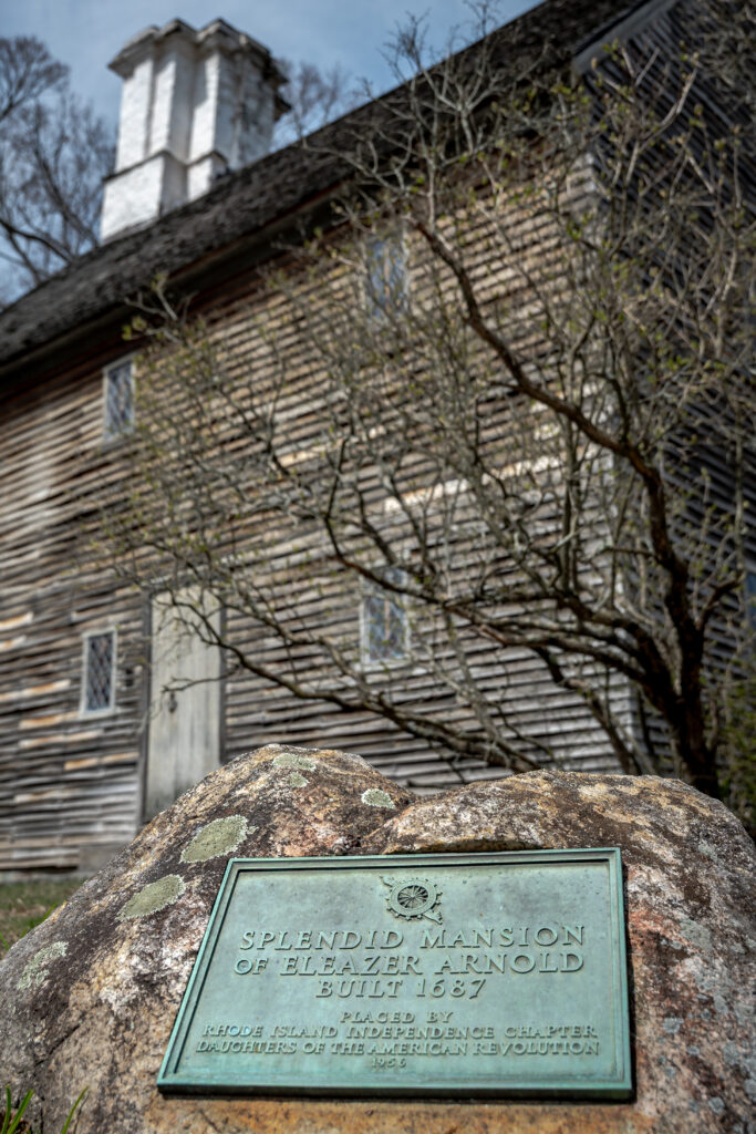 A historic wooden house with a white chimney and a plaque on a boulder in the foreground. Transcribed Text: SPLENDID MANSION OF ELEAZER ARNOLD BUILT 1687 PLACED BY RHODE ISLAND INDEPENDENCE CHAPTER DAUGHTERS OF THE AMERICAN REVOLUTION 1954