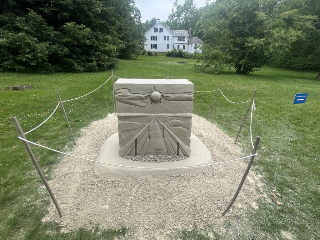 Sand sculpture with sun and triangle designs, surrounded by rope, with a house and trees in the background. Transcribed Text: "Please do not touch."