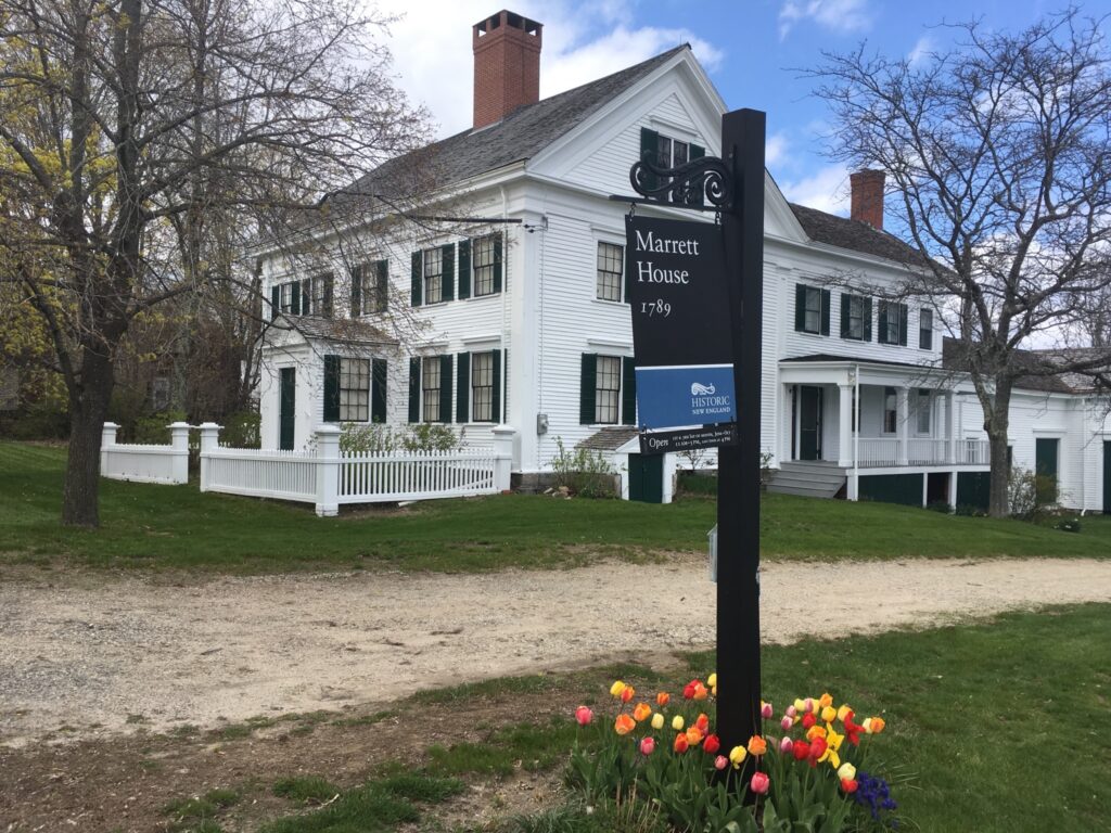 Historic white Marrett House with black shutters, picket fence, and a blooming tulip bed in front. Transcribed Text: Marrett House 1789 HISTORIC NEW ENGLAND Open