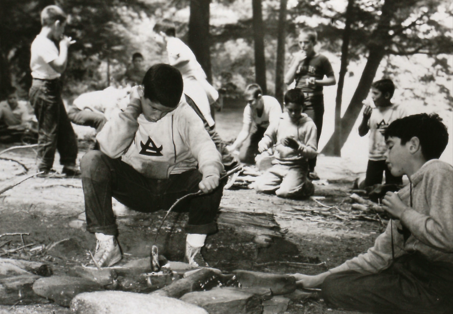 Boys in a wooded area, engaging in activities around a small fire.