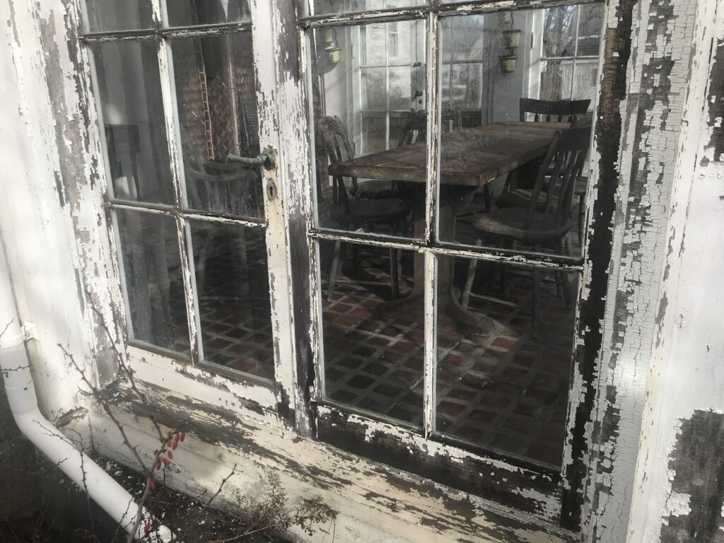 Weathered glass window with peeling paint, revealing a rustic interior with a wooden table and chairs.