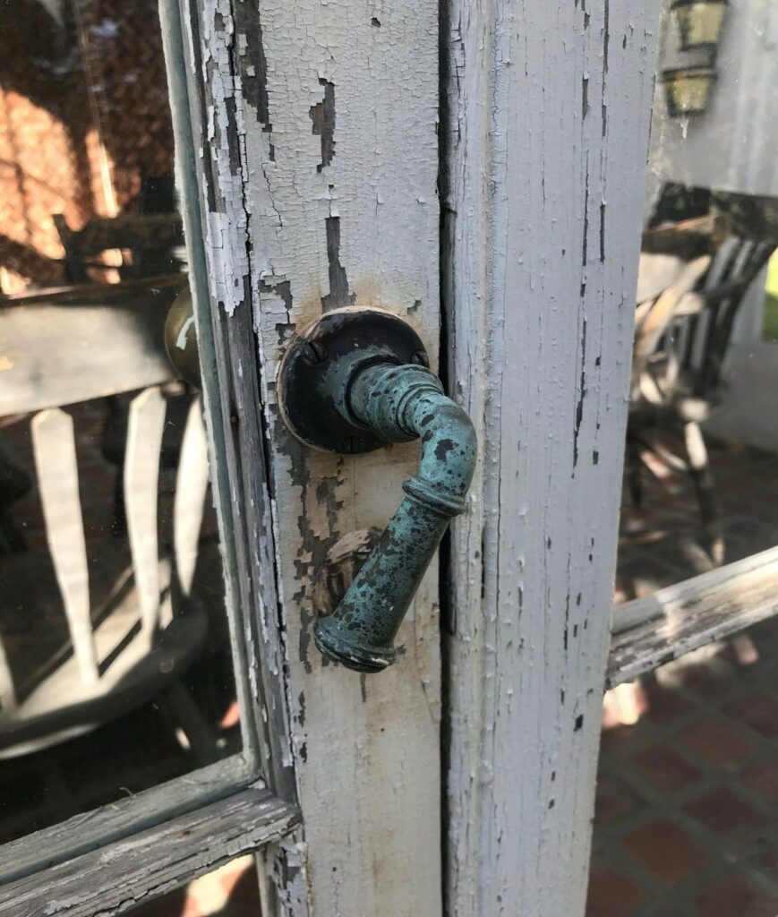 Close-up of a weathered wooden door with peeling white paint and a vintage metal handle with a greenish patina.