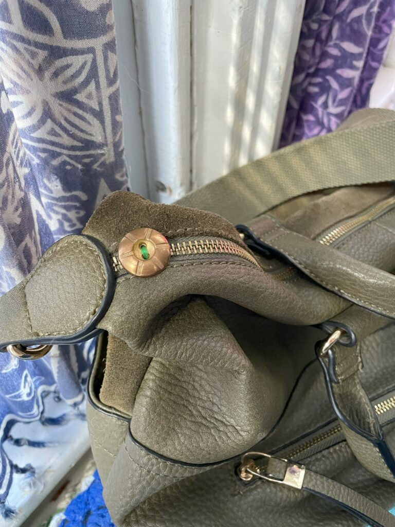 Close-up of an olive green leather bag with a decorative bronze button and patterned fabric in the background.