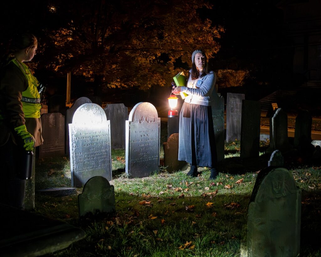 Two people in a cemetery at night, with headstones lit by artificial light.