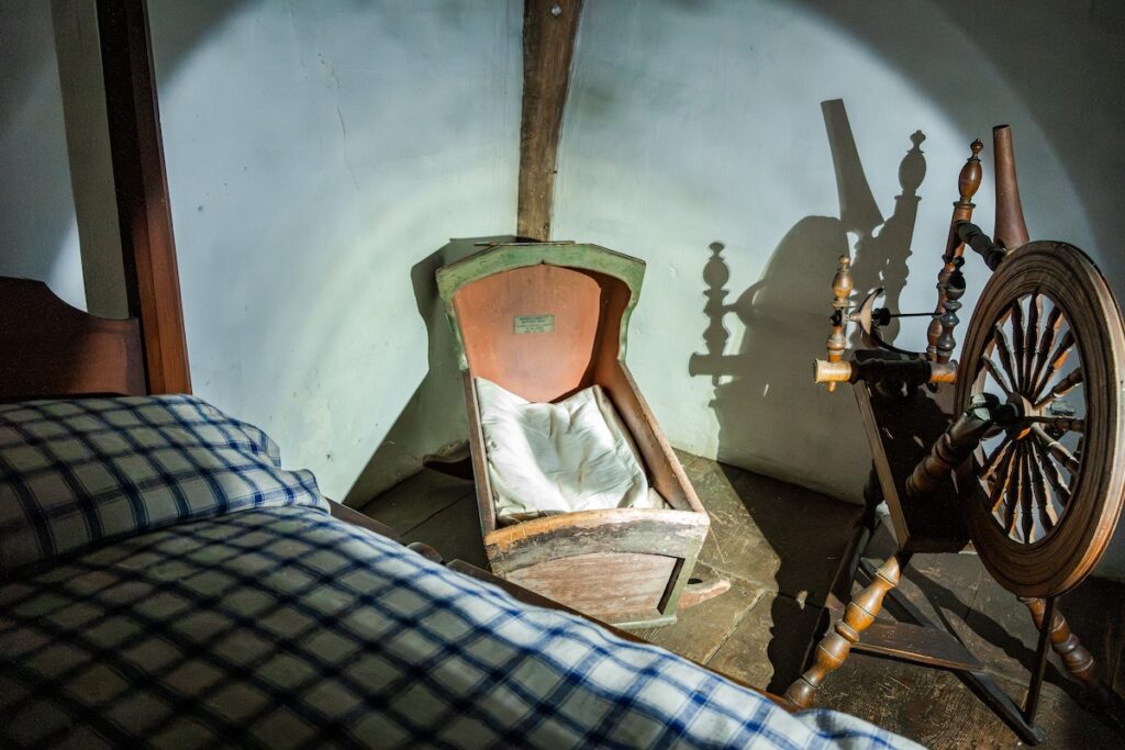 Antique wooden cradle with a white cloth inside, a checkered bed, and a spinning wheel in a rustic room.