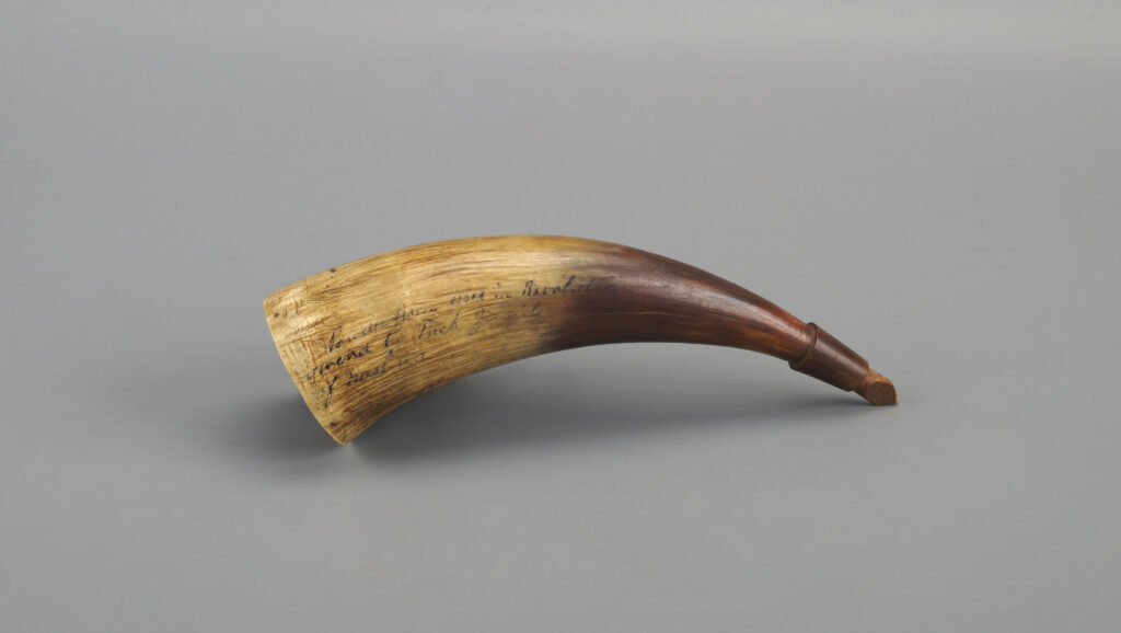 The image displays a powder horn with a natural curved structure, set against a neutral gray background. The horn exhibits a gradient of color from light tan at the wider end to a rich, dark brown near the narrow tip. The surface appears smooth, showing the natural texture of horn material. There is handwritten text visible on the lighter section of the horn, although it is partially faded and difficult to read. A small, cork-like stopper is inserted at the narrow end of the horn.