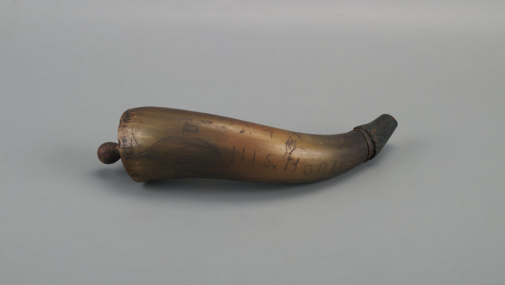 The image shows an antique powder horn laying horizontally against a neutral gray background. The horn is naturally curved, tapering to a narrow end, which appears darkened compared to the rest of the horn. The broader end features a wooden plug that is slightly protruding. The surface of the horn has a smooth texture and is light brown with darker engravings etched into it. These engravings include simple line drawings and text. The drawings illustrate a flag on a hill and a tree or plant shape. Various markings and scrawls can be seen across the horn, with the letters "HIS" and "MORR" visibly engraved.