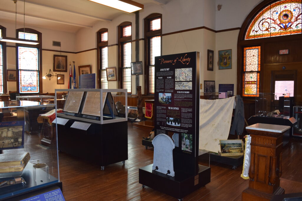 A museum room with stained-glass windows, display cases, and a "Pioneers of Liberty" panel.