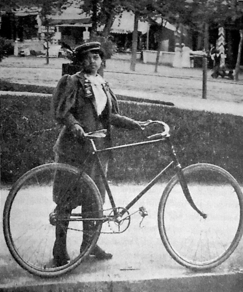 Person in early 20th-century attire standing with a vintage bicycle.