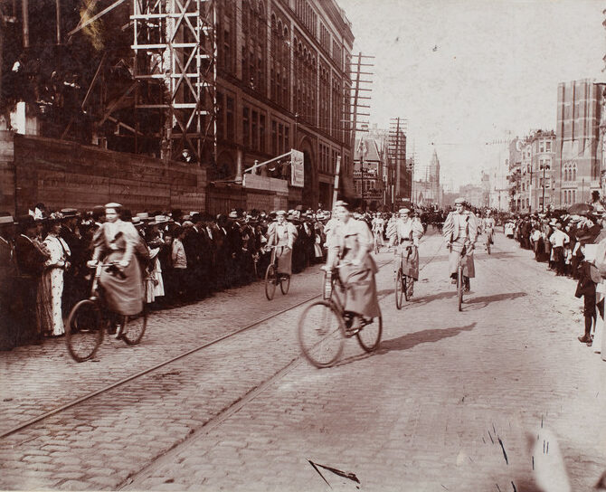 Historic street scene with cyclists on cobblestone road, surrounded by large crowds and old buildings.