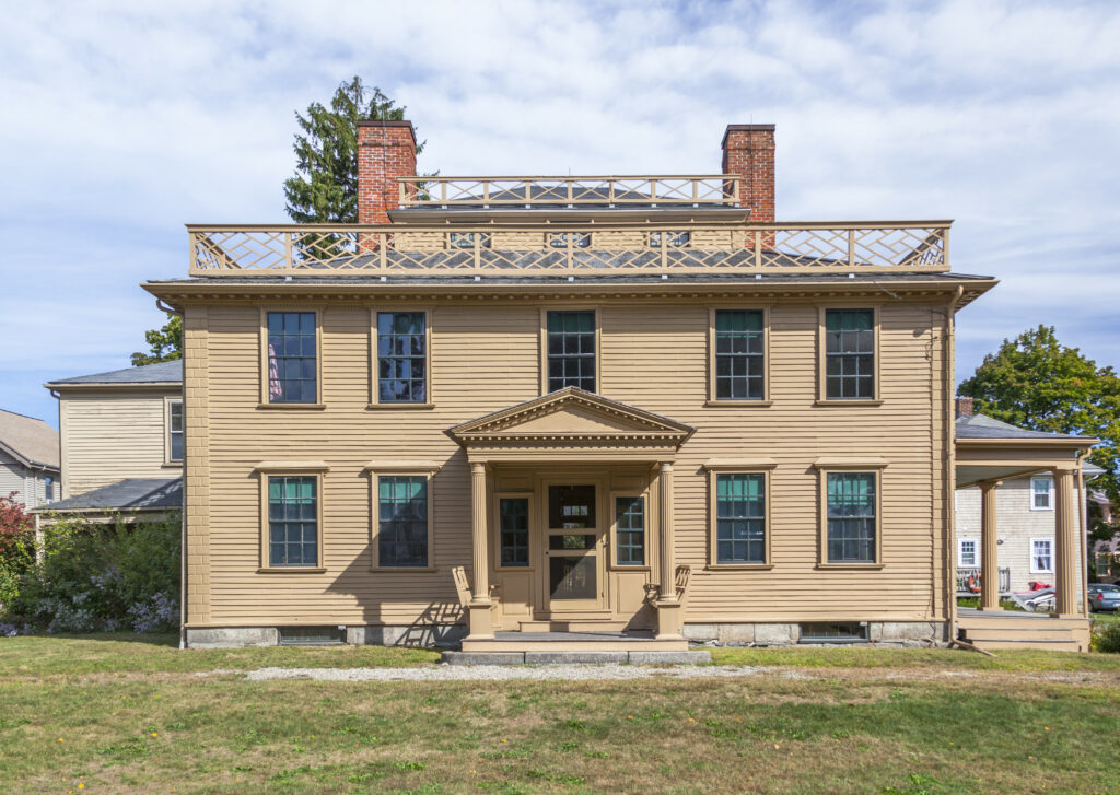 Classic two-story house painted beige with green-trimmed windows, a central porch, and brick chimneys.