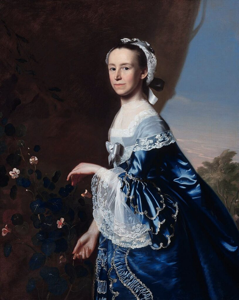 The image is a classical portrait of a woman standing in front of a dark, muted background. She is wearing an elaborate blue satin gown adorned with intricate lace details and gold embroidery. The dress features a white lace bodice, and her sleeves have ruffled lace edges. A white ribbon is tied into a bow at the front of her gown. The woman has fair skin, dark hair that is partly covered by a white lace cap, and she gazes directly at the viewer with a composed expression. Her hands are delicately positioned, with one slightly raised as if about to touch a flowering plant on the left, featuring dark leaves and small, pale pink blossoms. The background transitions on the right to a subtle suggestion of a landscape with a soft blue sky and distant greenery.