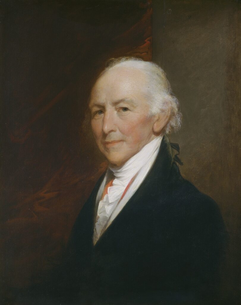 The image is a classic portrait of an older man with a balding head and white hair that curls at the sides. He is dressed in a formal manner, wearing a dark coat over a white shirt with high collar and intricate cravat. The background is a blend of dark shades, mostly brown and black, adding depth and focus to the man’s visage. The man's expression is calm with a slight, knowing smile, and his face is subtly lit, enhancing the contours and texture of his skin.