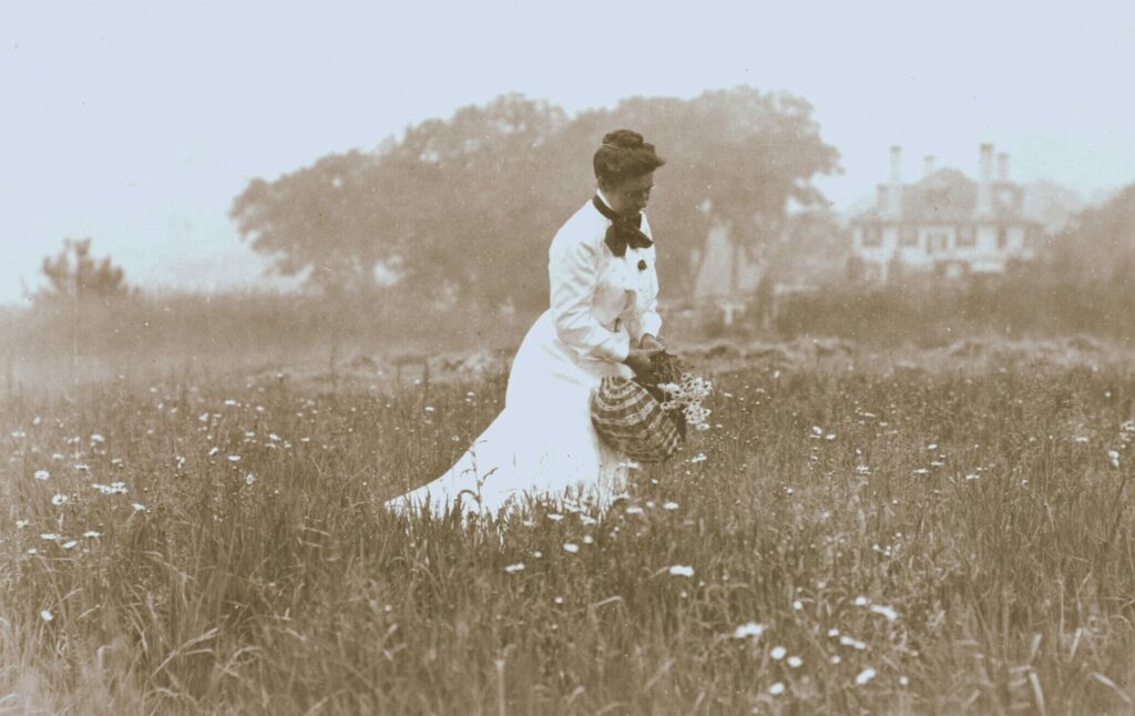 A sepia-tone photo of a woman in a field collecting flowers.