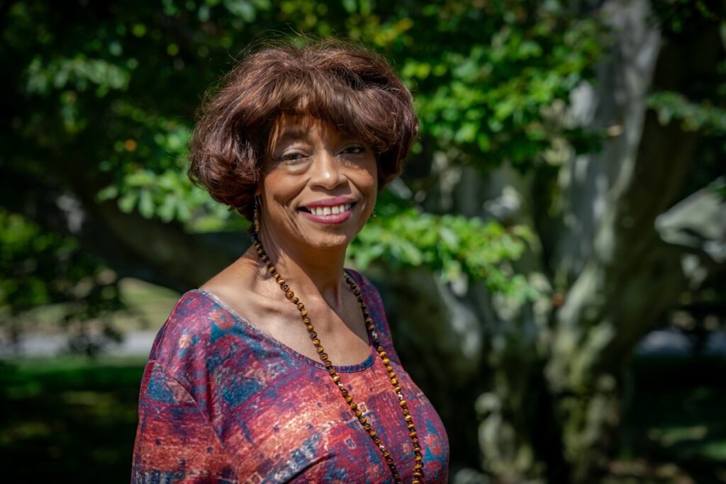 The image shows a person with short, wavy brown hair standing outdoors in front of a leafy tree. They are dressed in a colorful top featuring a pattern with various shades of red and blue. A long beaded necklace adorns their neck. The person is smiling and looking slightly toward the camera. Sunlight filters through the leaves, casting dappled shadows on them and the surrounding area. The background consists of lush green foliage and edges of tree trunks, with blurred hints of grass and pathways.