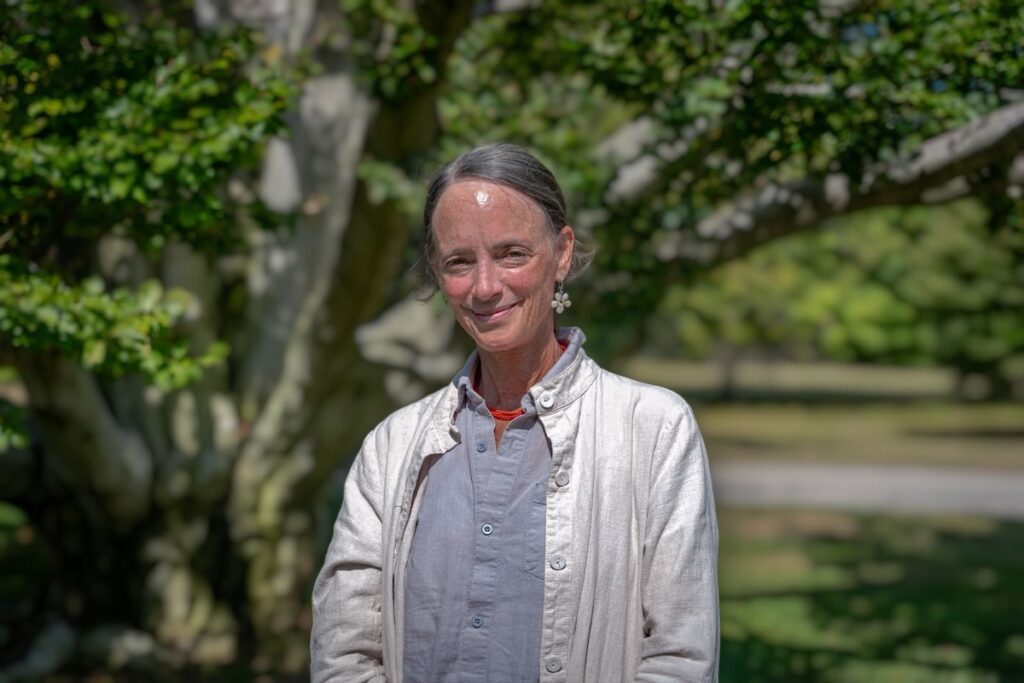 The image features a person standing outdoors with a soft smile. They are wearing a light-colored jacket over a dark shirt, and there is a red necklace visible. Their hair is neatly tied back, and they have earrings with a floral design. In the background, there is a large, mature tree with thick branches and lush green foliage. The lighting suggests it is a sunny day, as there are clear shadows.