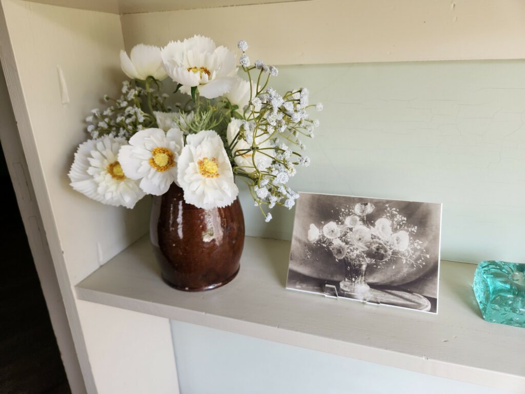 A vase with white flowers next to a black and white photo of similar flowers on a shelf.