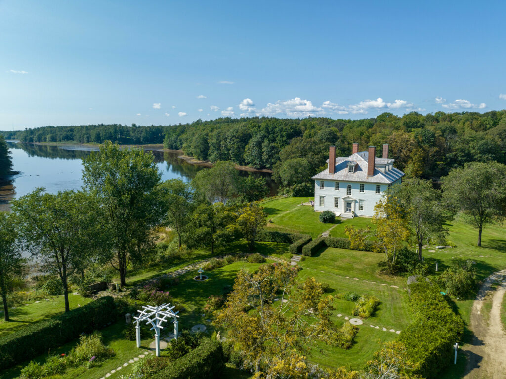 Aerial view of a white colonial-style house on a green lawn by a river, surrounded by gardens and trees.