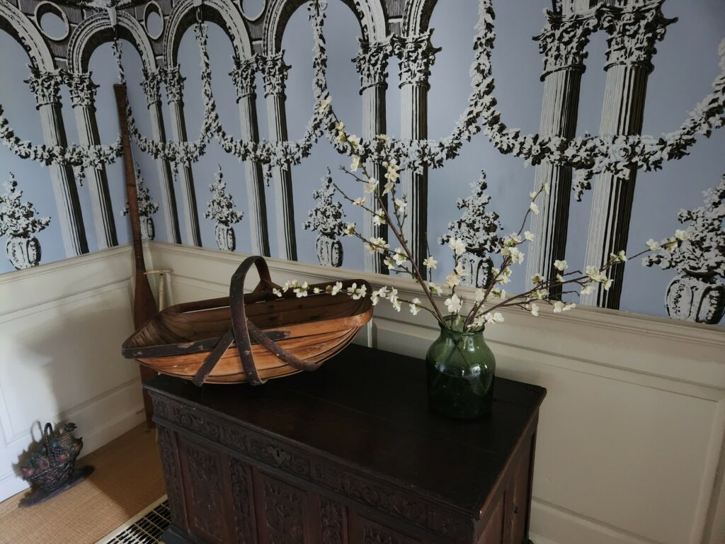 Room corner with classical wallpaper, a wooden cabinet, a vase with white flowers, and a wooden bowl.