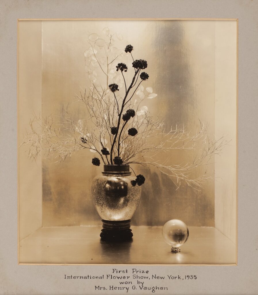 Sepia-toned photo of a floral arrangement in a reflective vase with a glass ornament beside it.