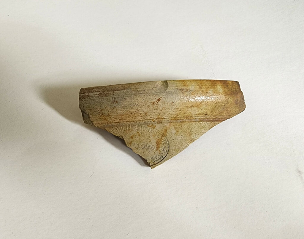 A fragment of pottery with earthy tones and faint markings, placed on an off-white background.