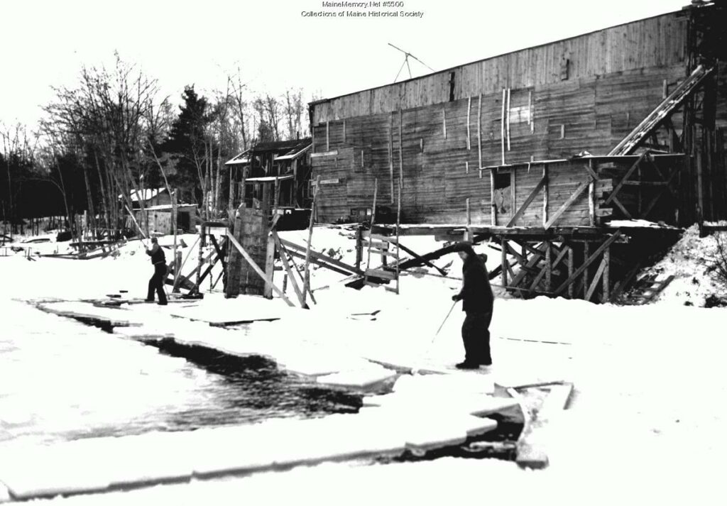 Two individuals harvesting ice on a frozen body of water near a large wooden structure. Transcribed Text: Mainememory.net #9550 Collections of Maine Historical Society