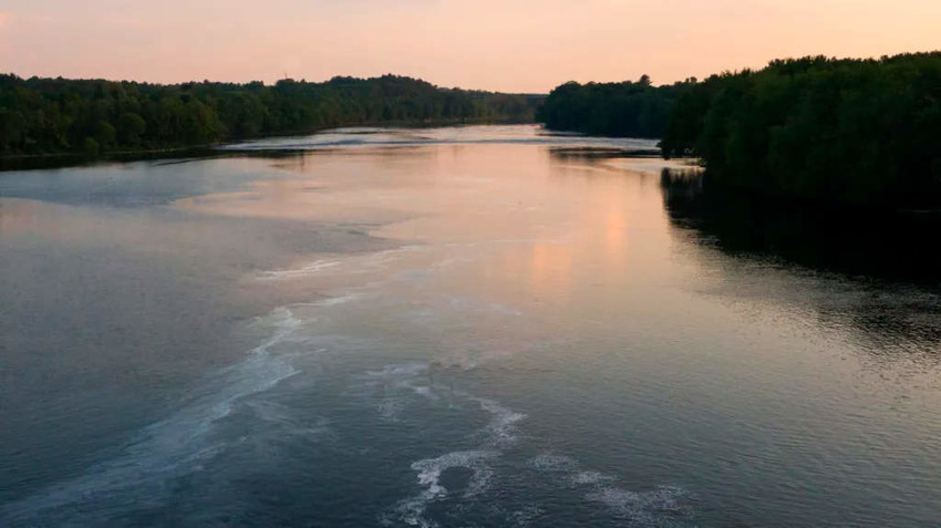 A wide river at sunset with reflections of the sky and bordered by dense green trees.