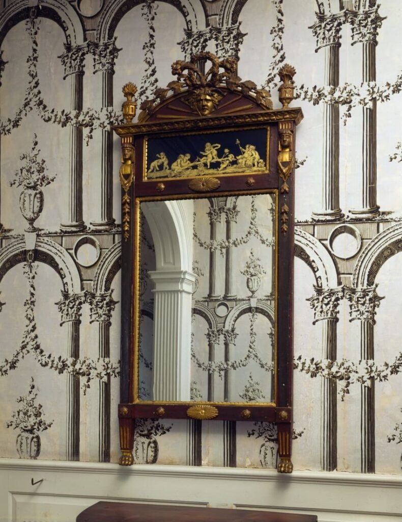 Ornate mirror with classical figures relief on wallpapered wall with arches and columns.