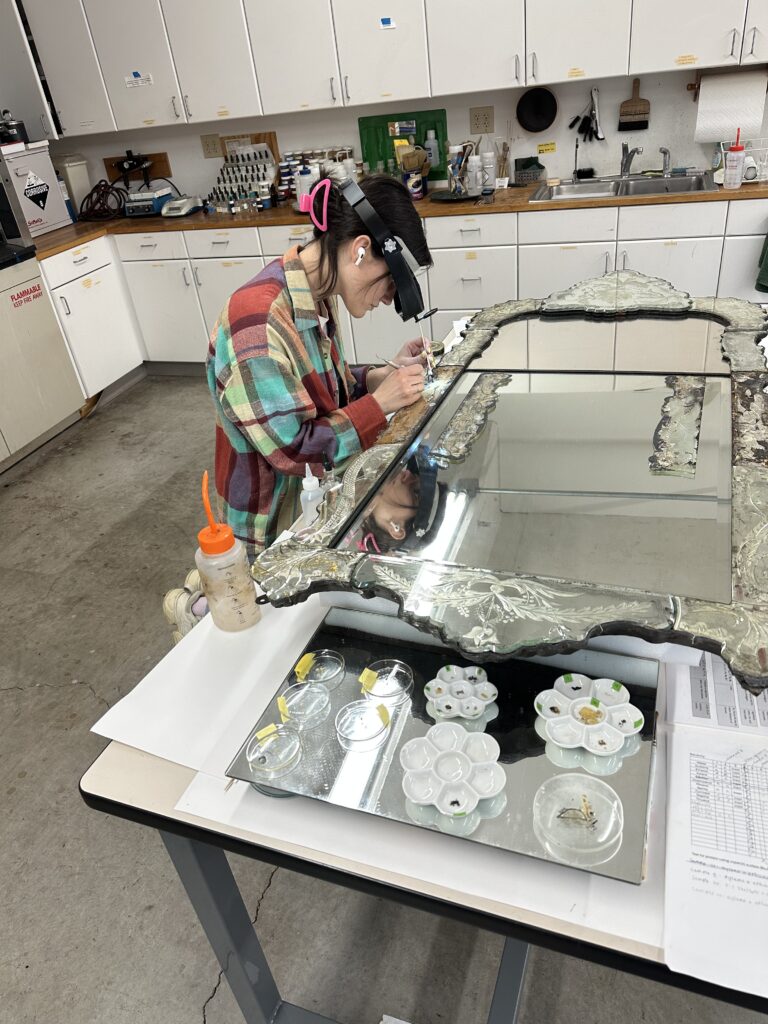 A woman in a workshop working on an ornate mirror with art supplies on the table.