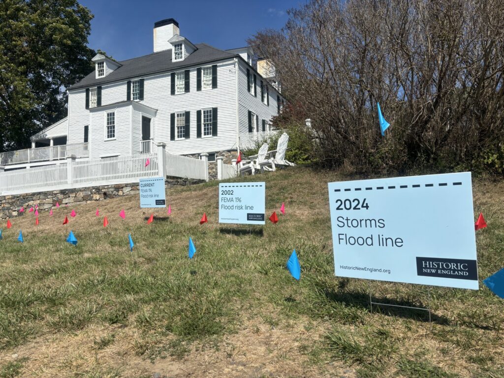 A white house on a grassy slope with signs marking flood risk lines and colorful flag markers. Transcribed Text: CURRENT FEMA 1% Flood risk line 2002 FEMA 1% Flood risk line 2024 Storms Flood line HistoricNewEngland.org